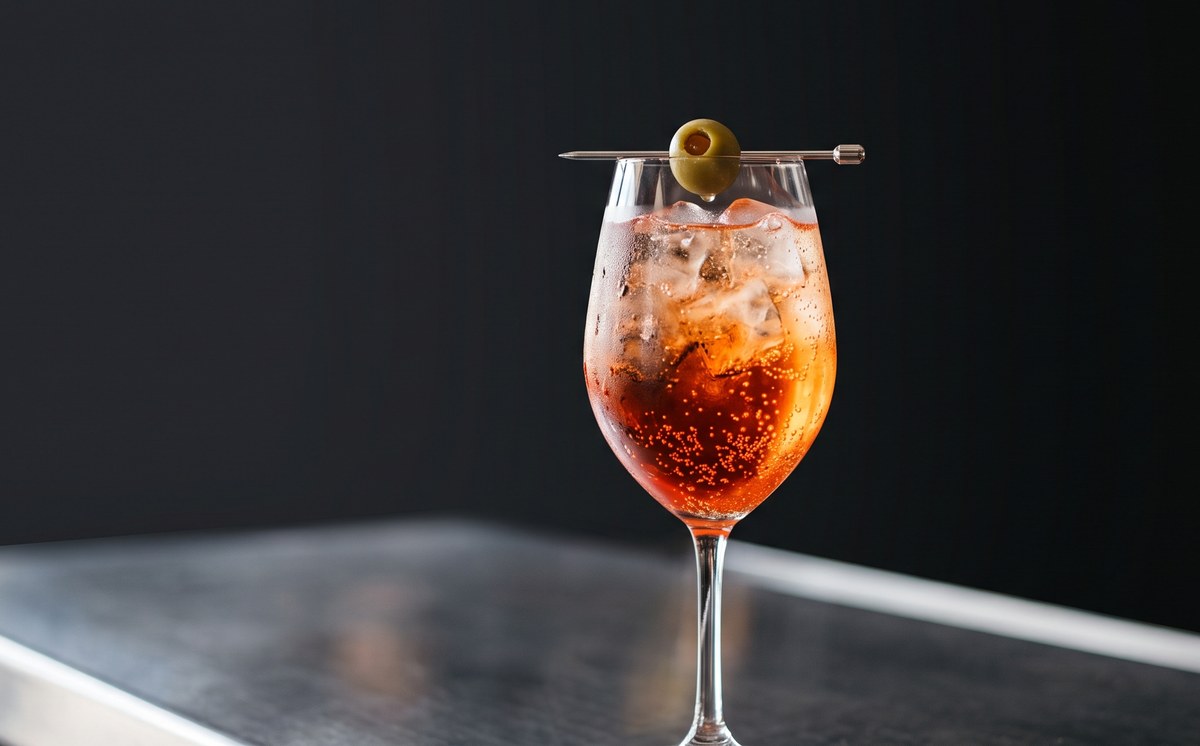The Lacto Pepper Spritz in a stemmed wine glass full of clear ice, deep orange-red and carbonated, a green olive on a pick across the rim, on a polished stainless bar top