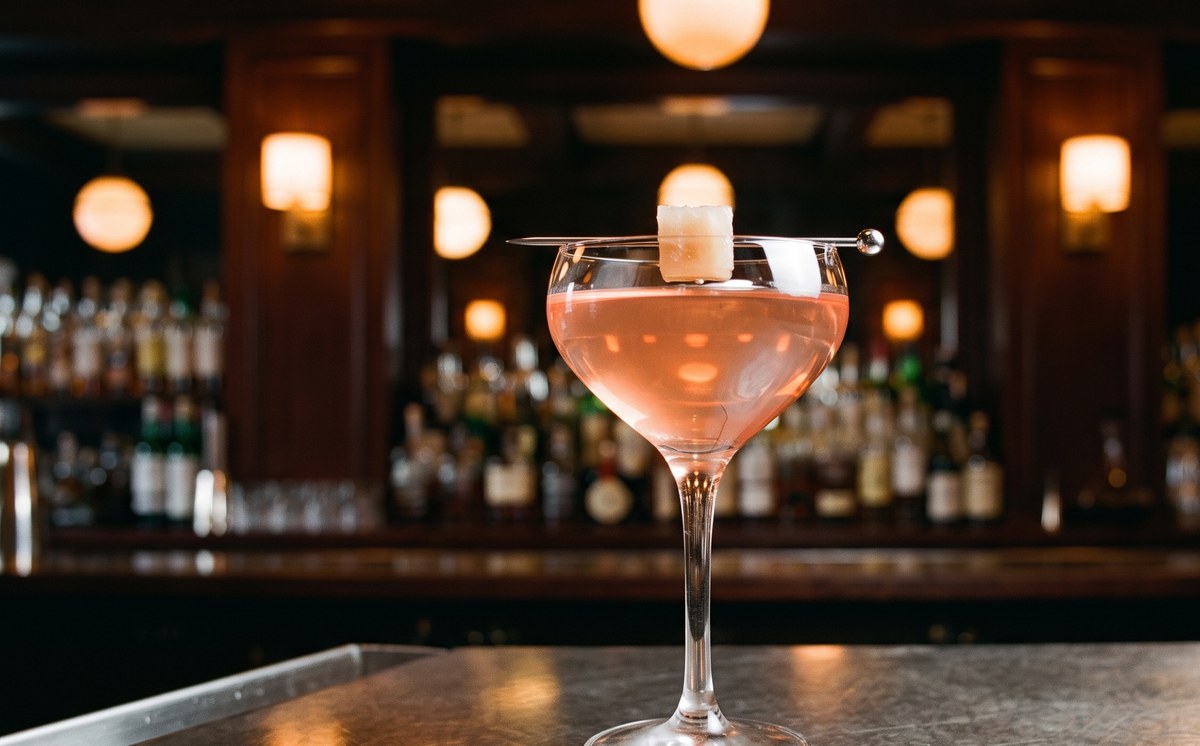 Watermelon rind gimlet served up in a chilled Nick and Nora glass, clear pale coral, one cube of pickled rind on a pick across the rim